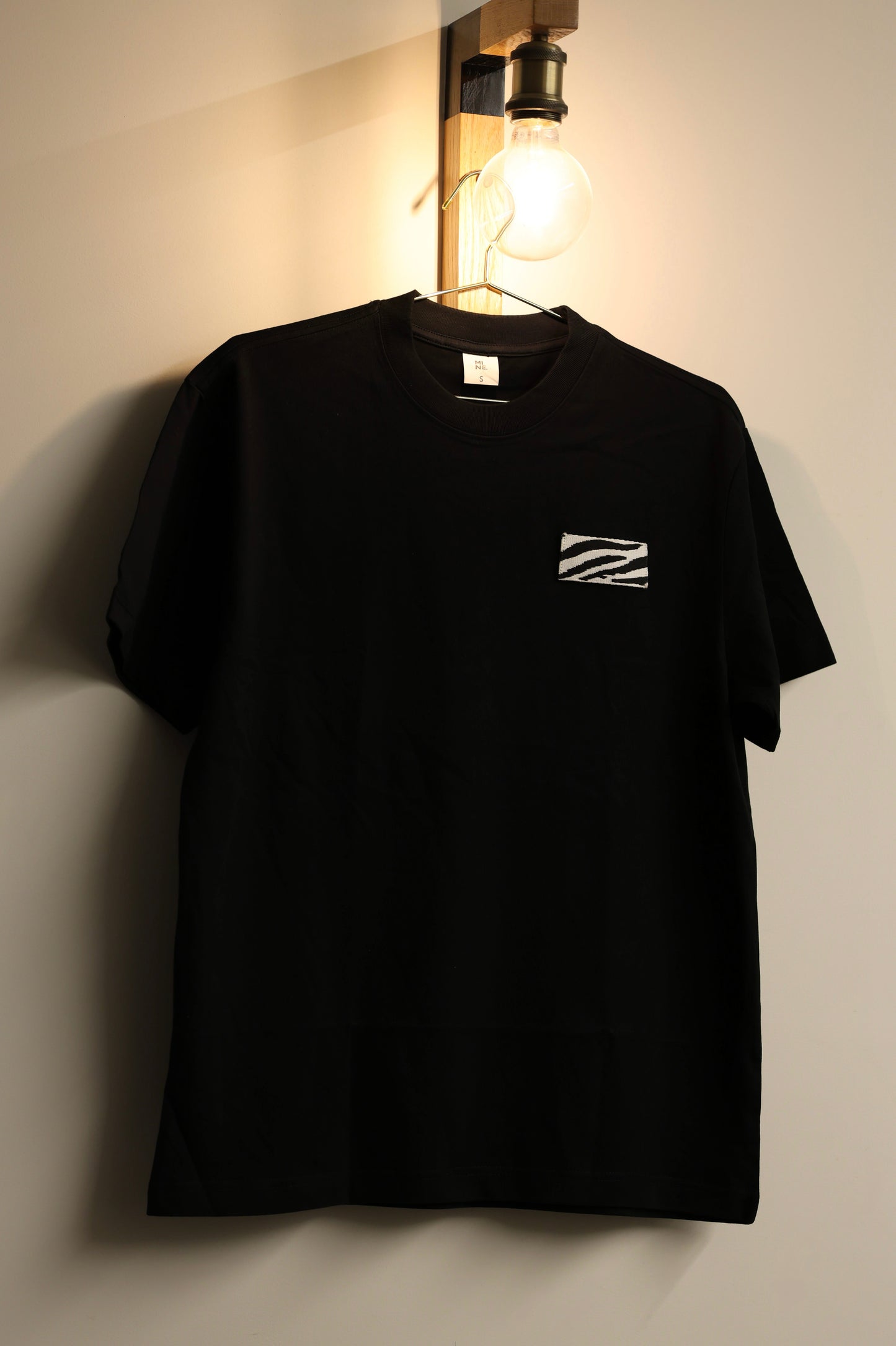 Black t-shirt with a small white logo on a light-colored wall.