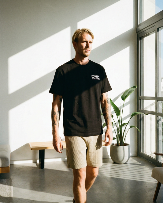 Man wearing a black t-shirt with a logo in a bright room with large windows and a plant.